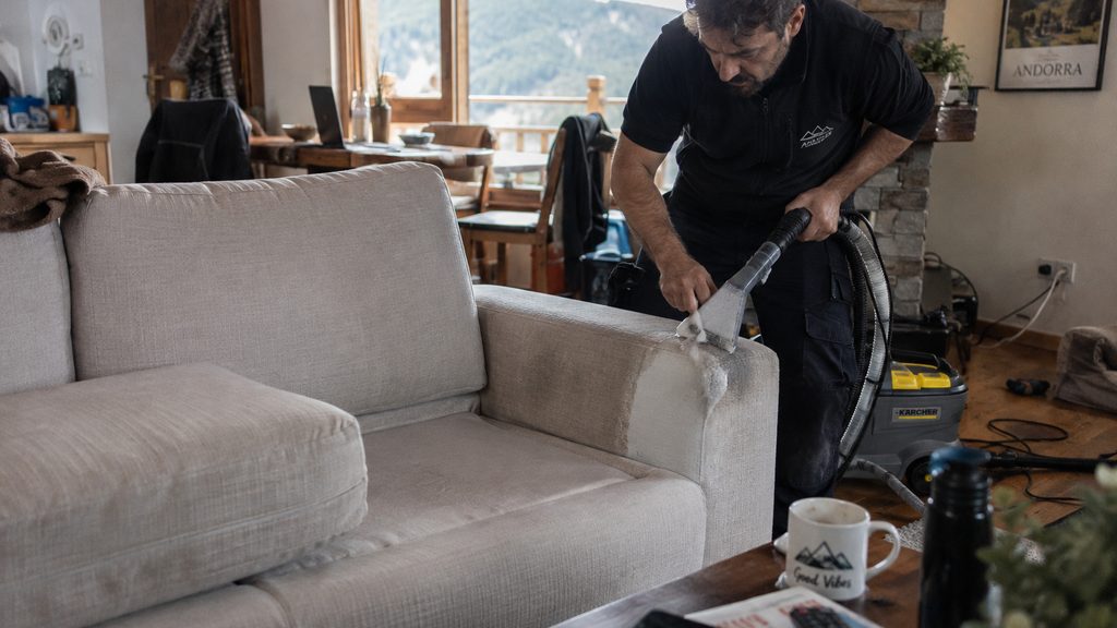 Operario limpiando el brazo de un sofá de tela en un salón de Andorra con media parte ya tratada